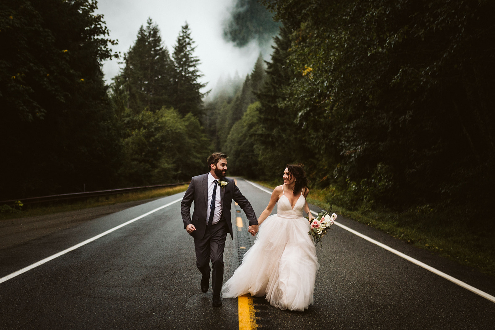 A bride and groom run hand-in-hand down a road surrounded with misty evergreen woods.