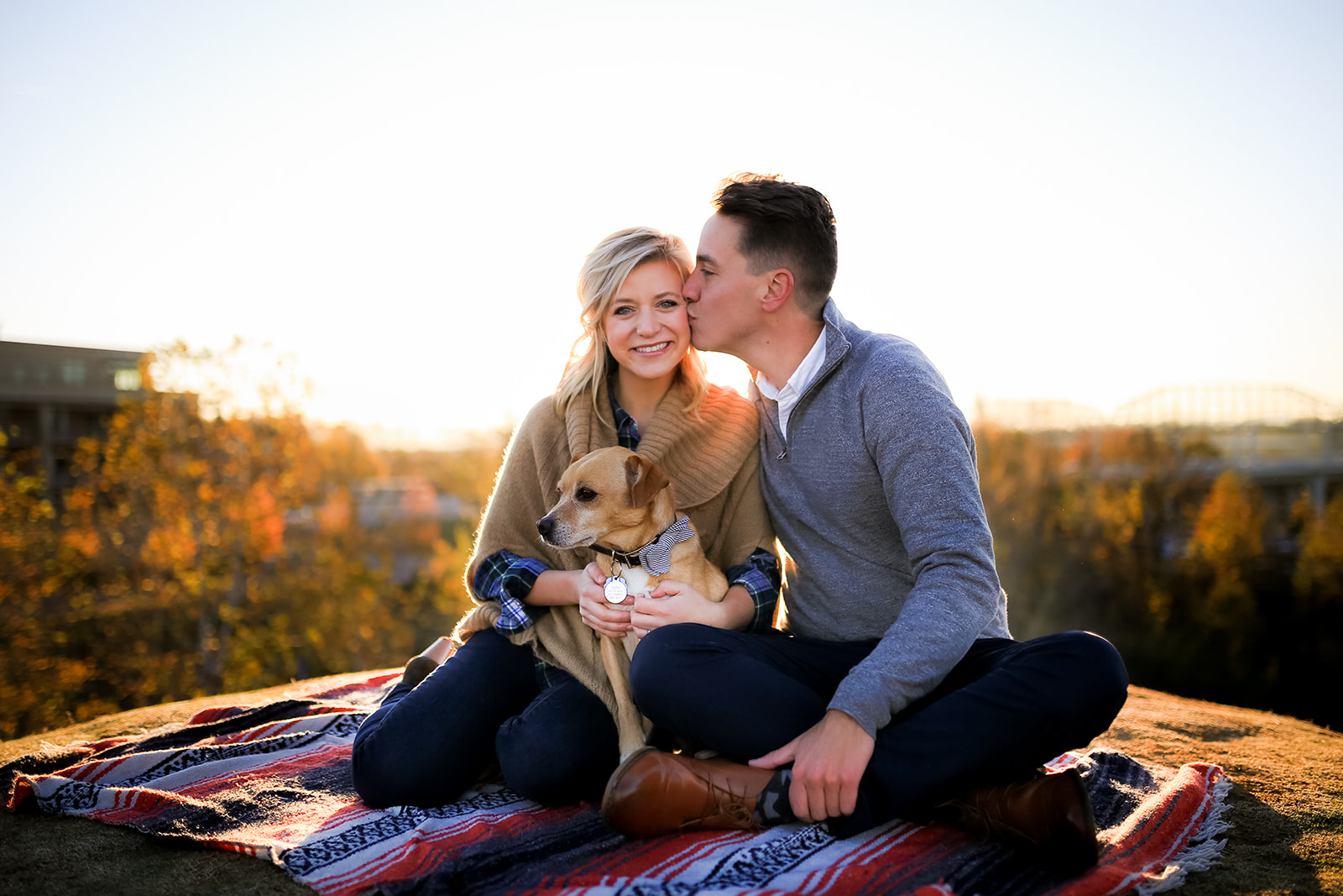 A couple cuddles their dog on a picnic blanket. The man kisses the woman's cheek as the sun sets behind them.