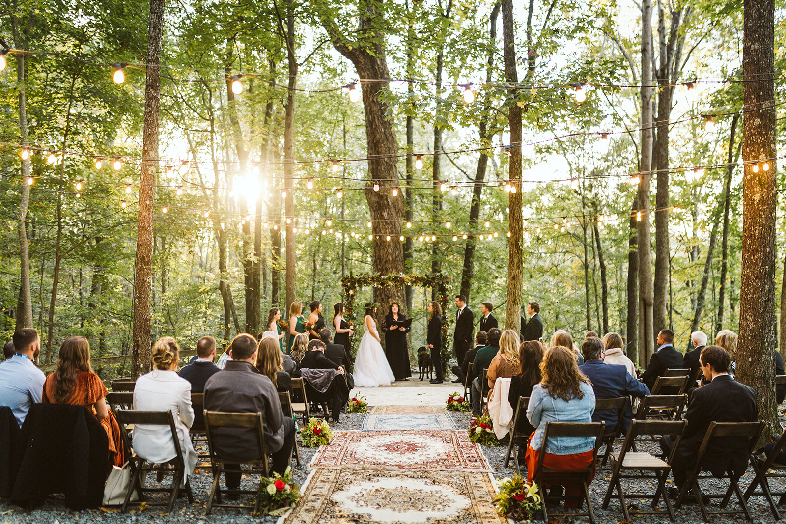 Bride and groom exchange vows in a forest as friends and family look on. The aisle is lined with vintage rugs, and the altar is lit up with strings of lights.