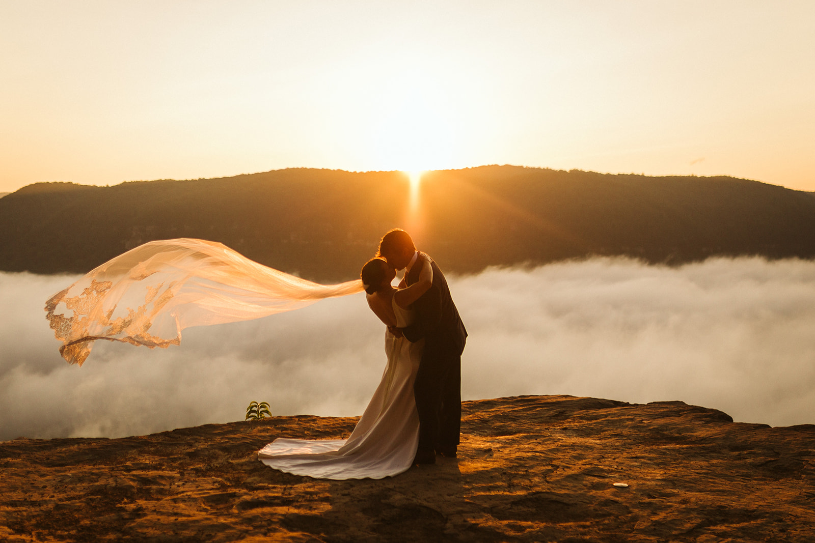 A bride and groom kiss under the setting sun on a mountain ridge. Her veil blows behind her in the wind.