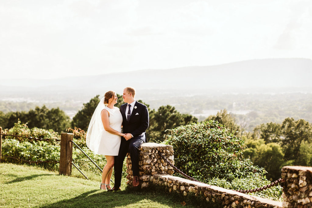 A bride and groom stand together and admire one another next to a short stone wall.