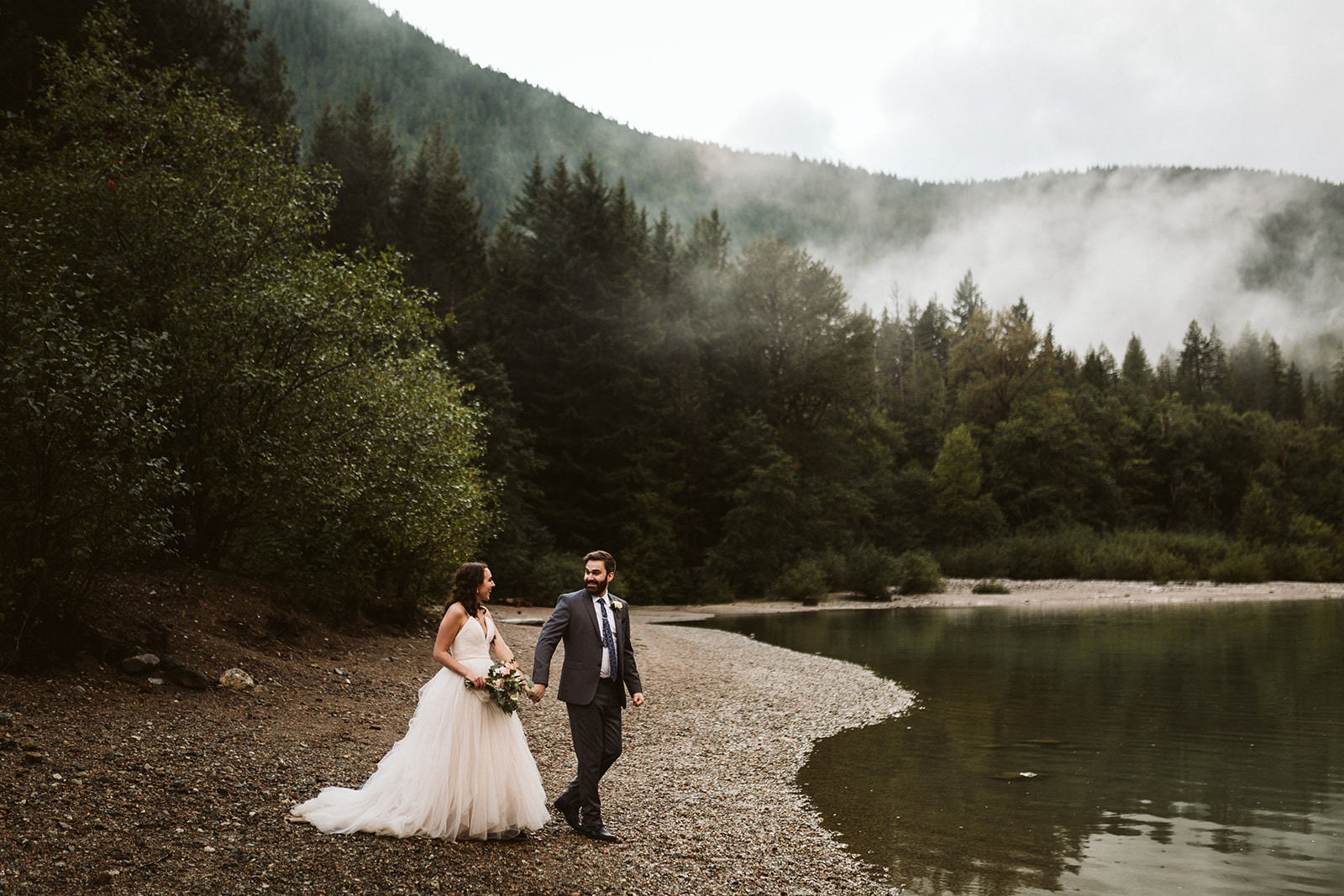 Bride and groom walk hand in hand on a rocky riverbank.