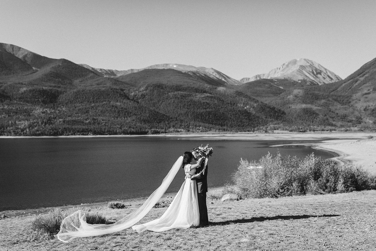 A bride and groom embrace in front of a mountain lake.