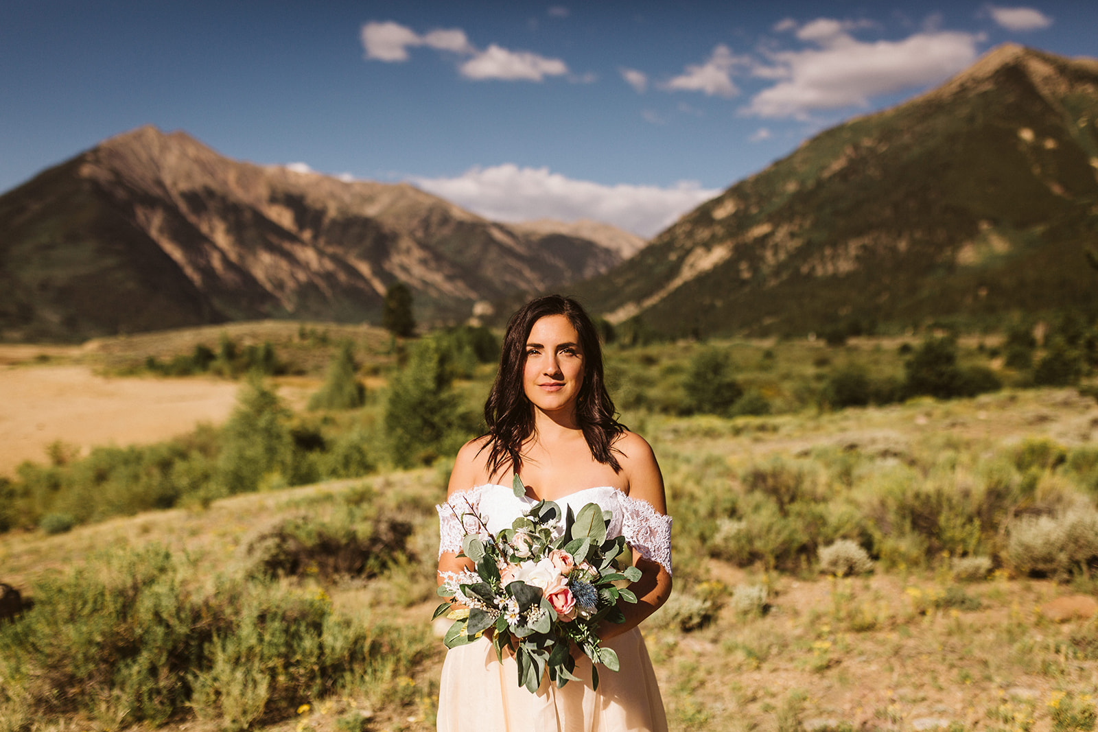 A bride poses straight-faced with her bouquet in front of green mountains.