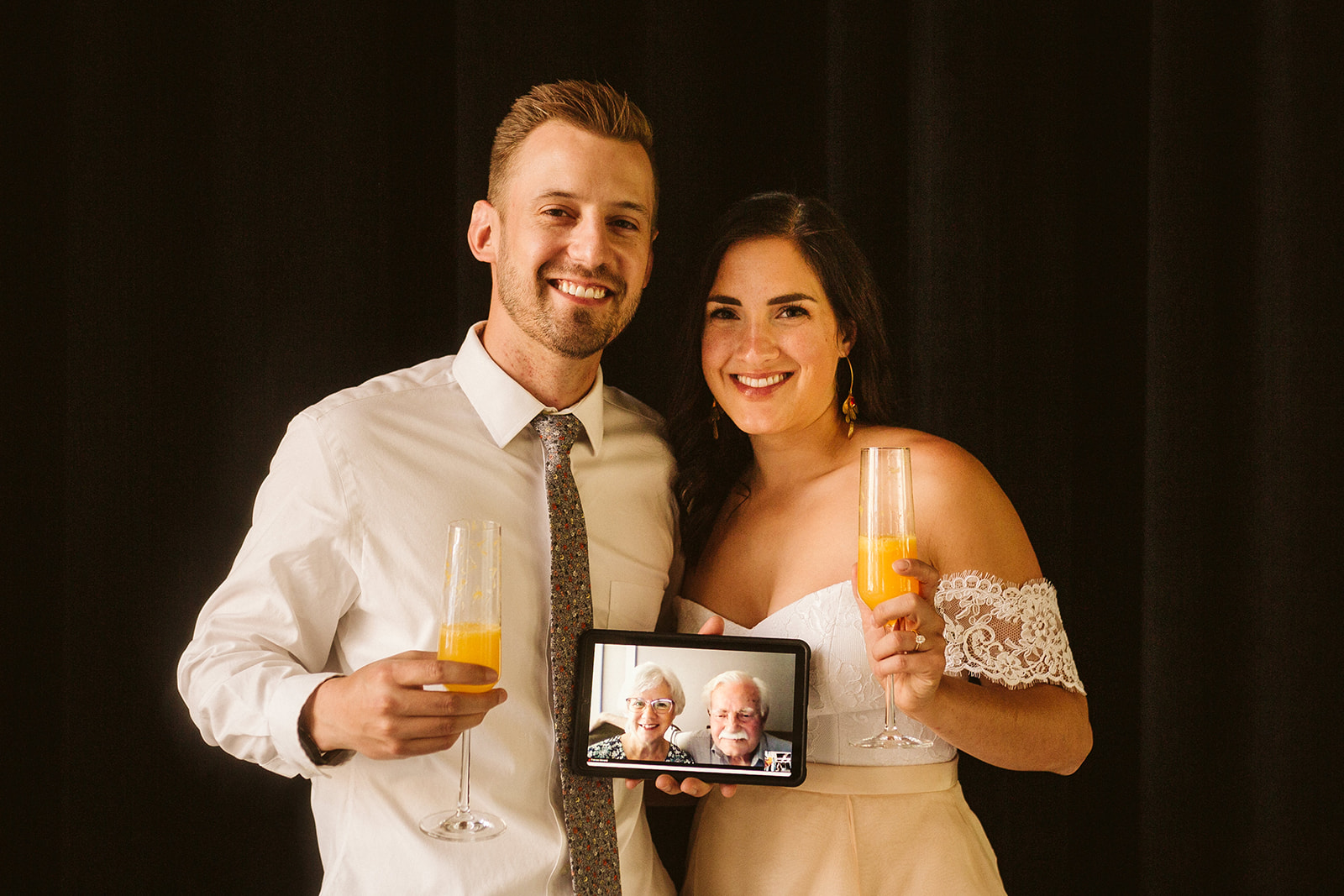 A bride and groom pose with mimosas while holding an iPad, which they're using to Facetime with family.
