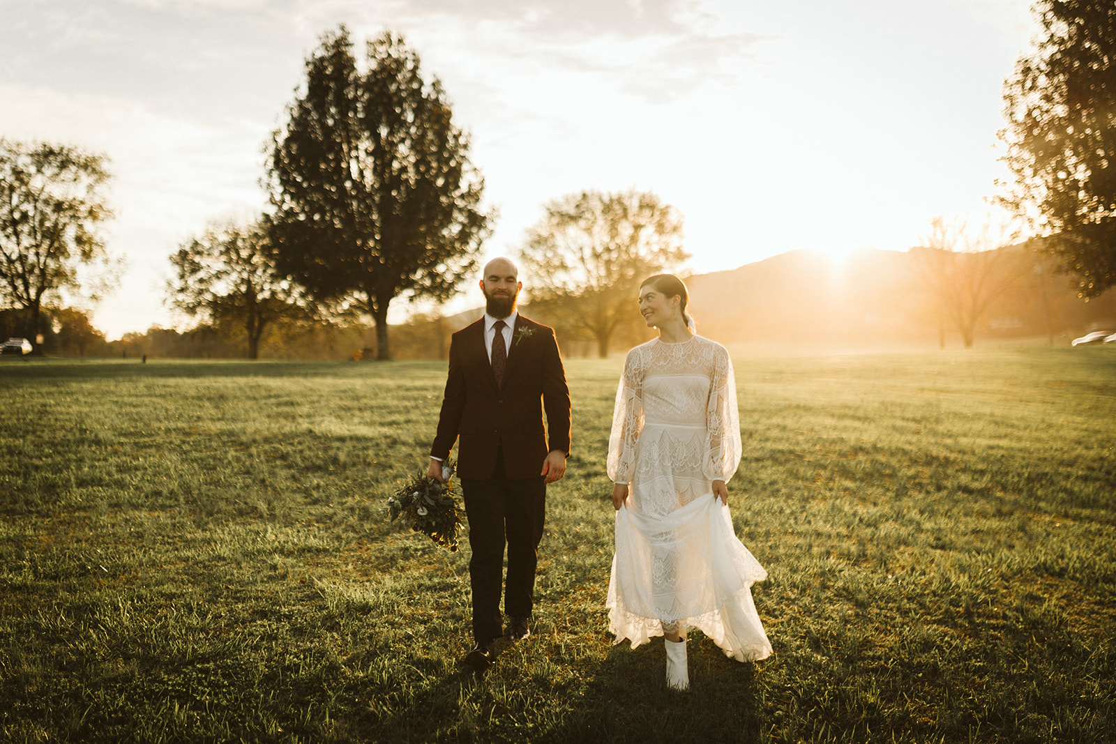 Bride holds her dress and smiles at the groom as they walk through the field at the Tennessee Riverplace