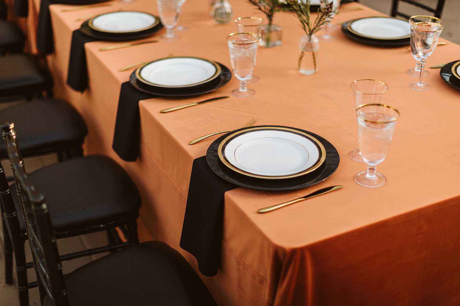 Close up of the gold silverware and gold rimmed plates with gold rimmed glasses sit on an orange cloth table