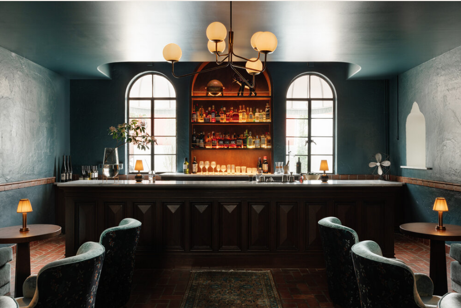 The cocktail lounge inside The Common House. The walls are painted dark teal, and the bottles behind the bar are illuminated in amber light.