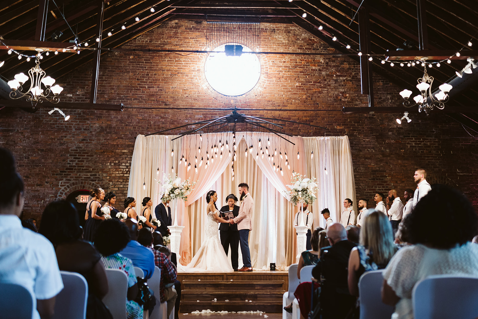 A bride and groom exchange vows in front of white drapery on an exposed brick wall.