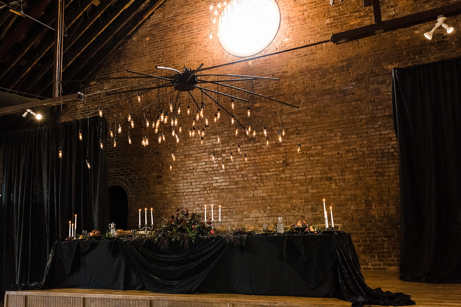 The head table at a wedding decorated in dark, gothic drapery and florals. There is a modern-looking light fixture suspended above the table.