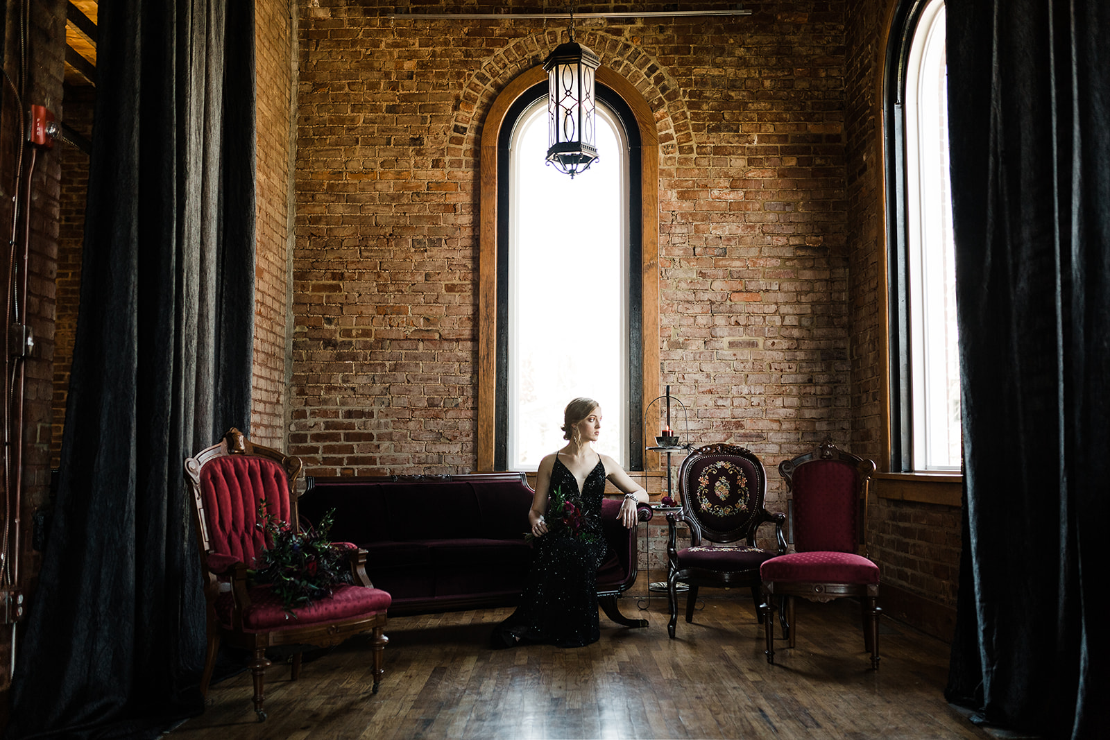 A bride in a black dress poses on a red velvet couch in front of an exposed brick wall with a tall, arched window.