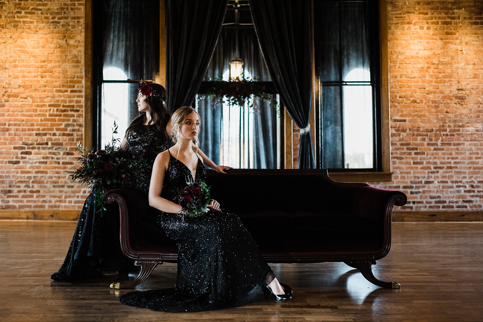 Two women in black dresses pose on a red velvet couch in a modern wedding venue.