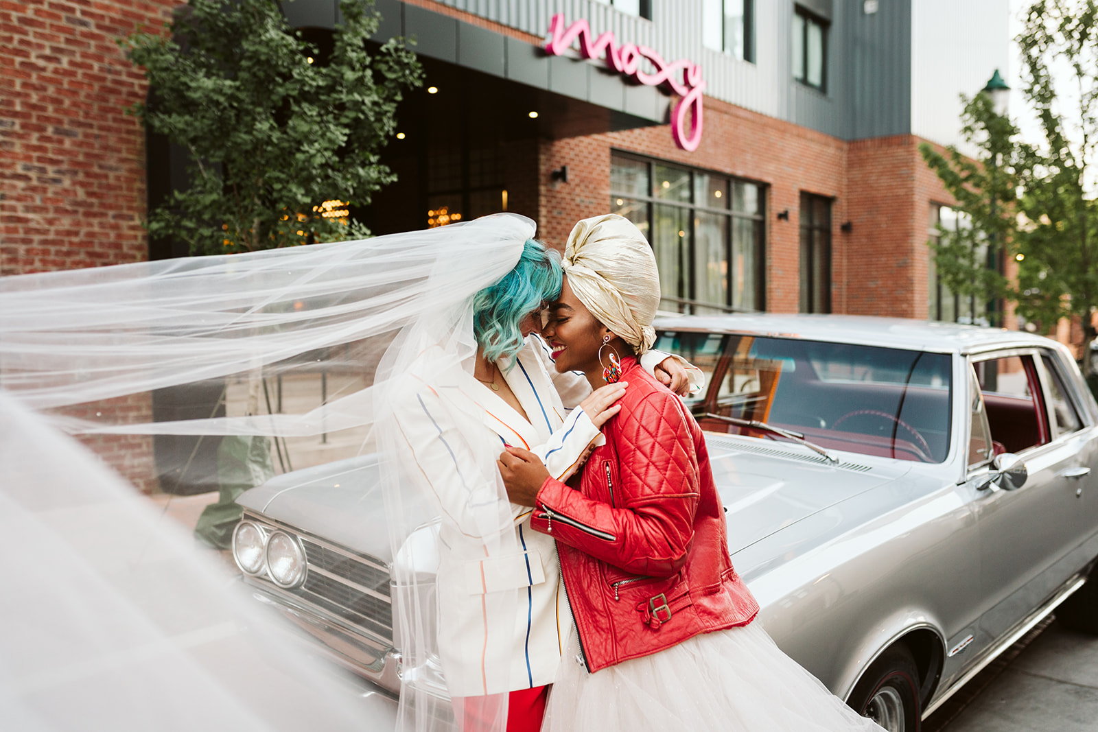 The brides lean in for a kiss in front of a classic car outside Moxy Chattanooga, a modern wedding venue.