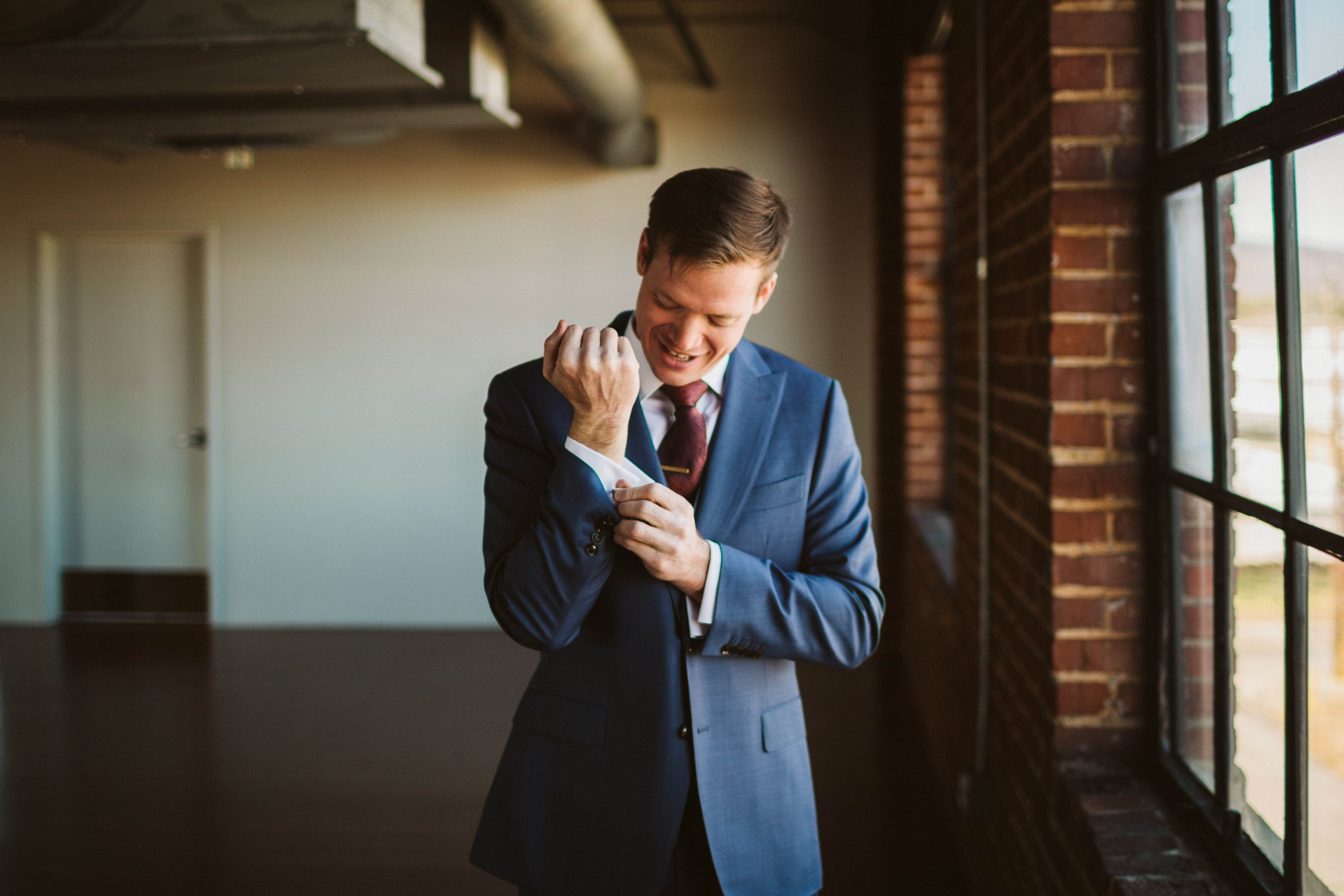 The groom fixes his cufflinks in front of a large sunny window.