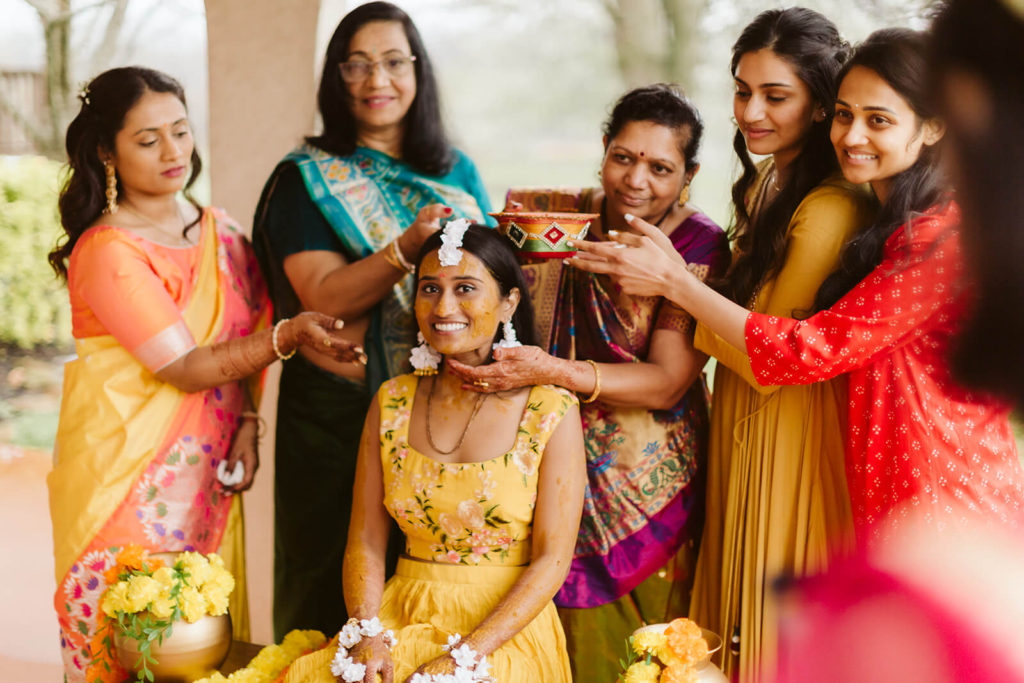 Bride's family members appling tumeric paste to her during Pithi ceremony. Photo by OkCrowe Photography.