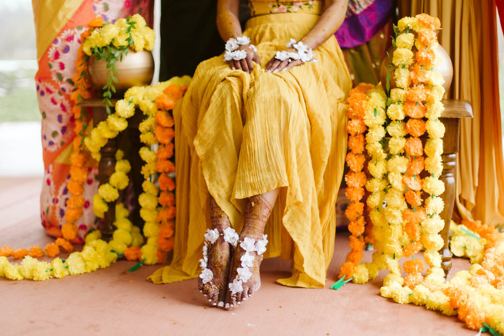 Indian bride in a yellow dress seated on a chair covered in yellow and orange flowers. Photo by OkCrowe Photography.