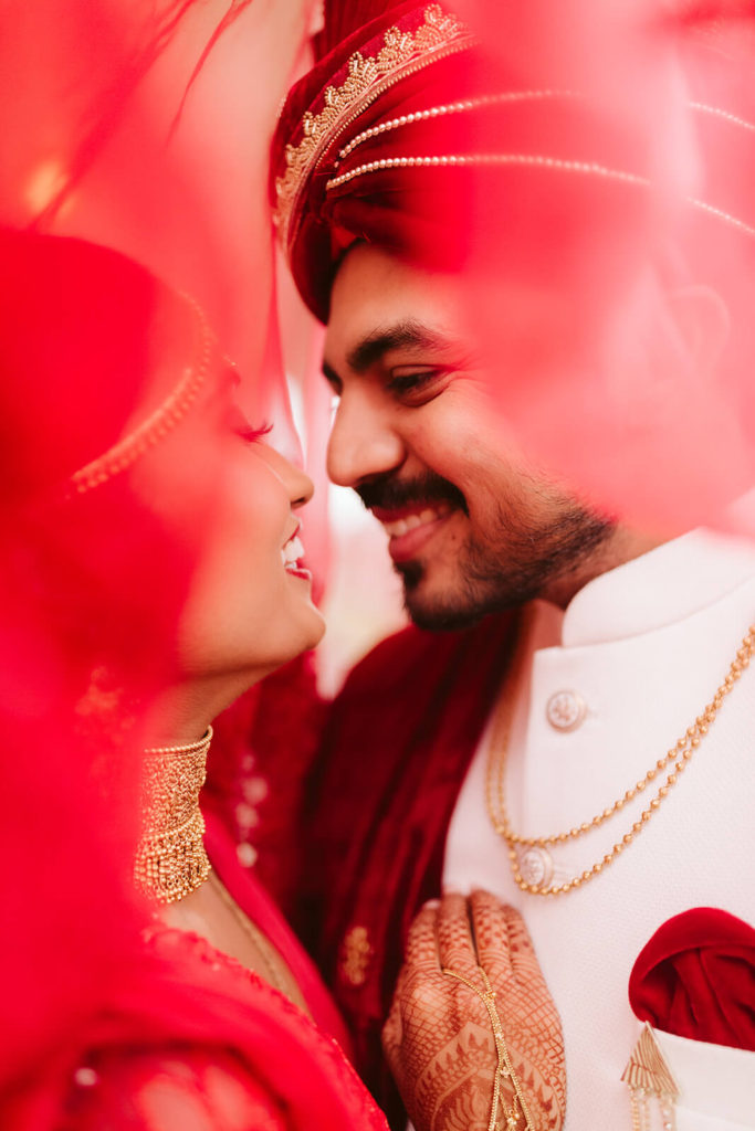 Indian bride and groom posing beneath her red veil. Photo by OkCrowe Photography.