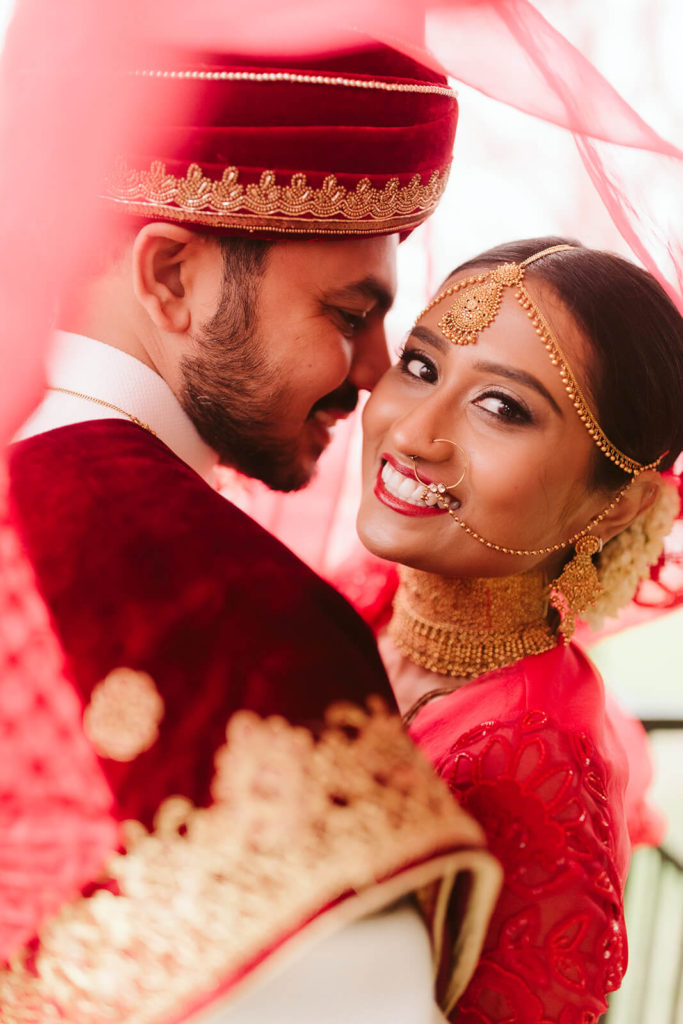 Indian bride and groom posing together beneath her red veil. Photo by OkCrowe Photography.