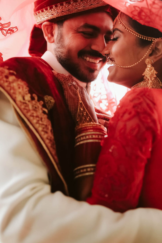 Bride and groom wearing traditional Indian wedding garments laughin beneath her red veil. Photo by OkCrowe Photography.