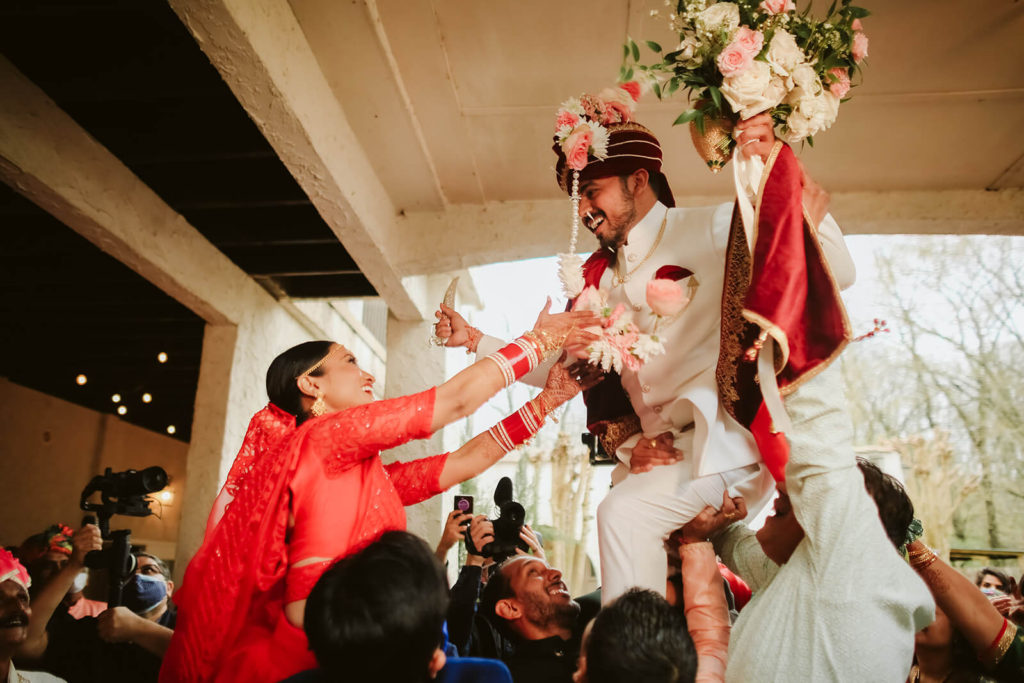 Indian groom arriving at the wedding venue surrounded by guests as his bride welcomes him. Photo by OkCrowe Photography.