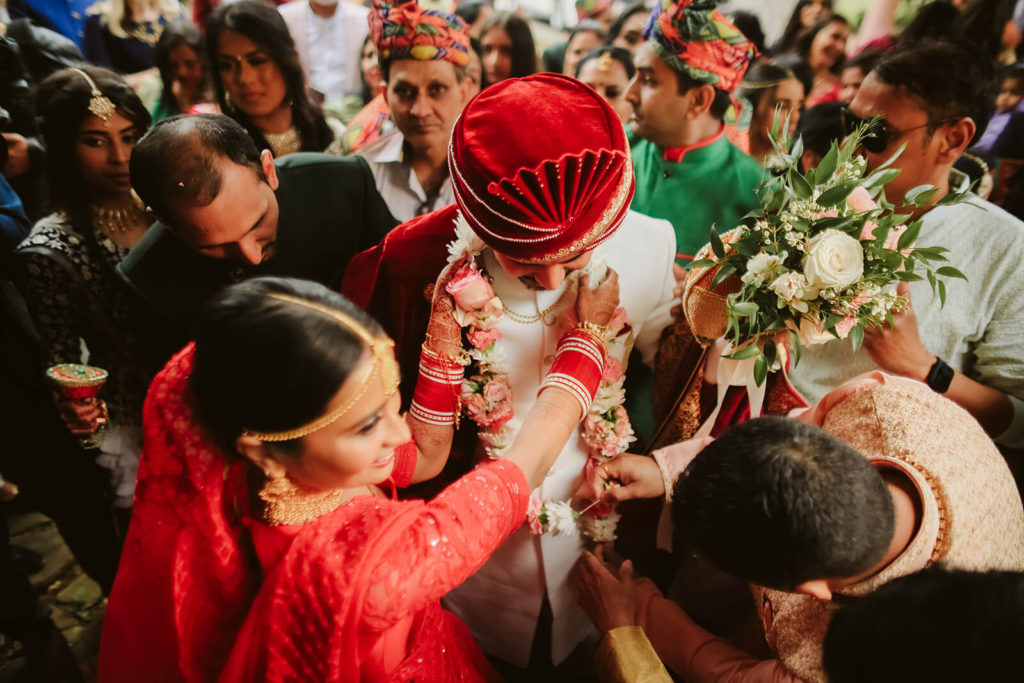 Bride placing a flower garland around groom surrounded by guests. Photo by OkCrowe Photography.