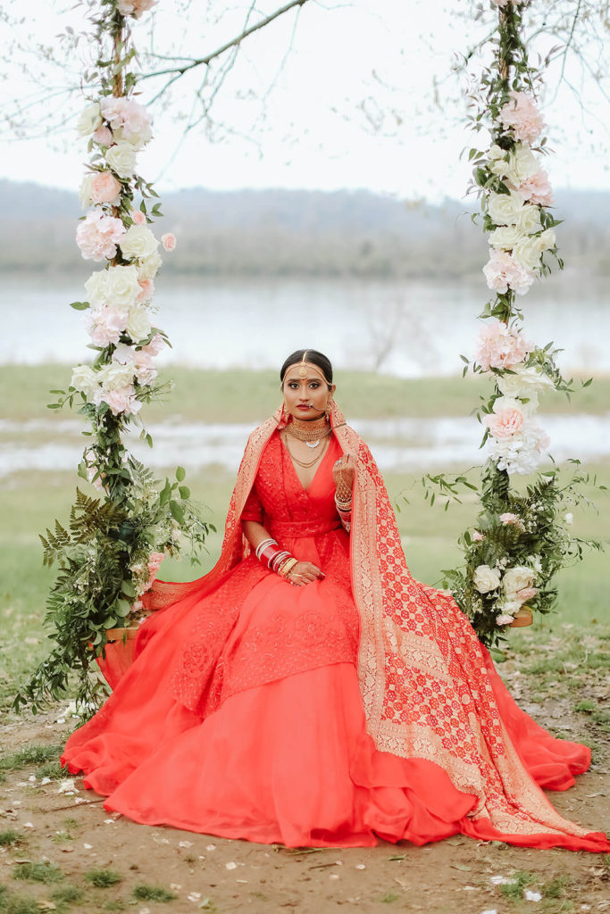 South Asian bride in traditional red wedding dress with red and gold shawl seated on a floral swing. Photo by OkCrowe Photography.