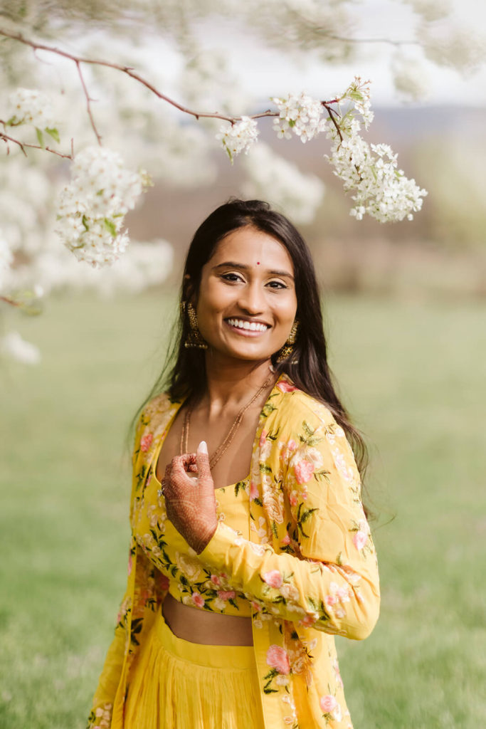 Bride wearing a traditional South Asian yellow dress under a white flowered tree. Photo by OkCrowe Photography.